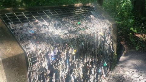 A large grate where the front is encrusted with detritus, including many wipes and cloths.