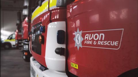 Photo showing the front bumper and bonnets of a line of fire engines, lined up inside a station. The one nearest to the camera has "Avon Fire and Rescue" in white writing on a red panel