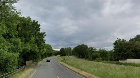 The A24 is seen, a dual carriageway with two lanes either side and a grass verge in the middle. There are trees either side