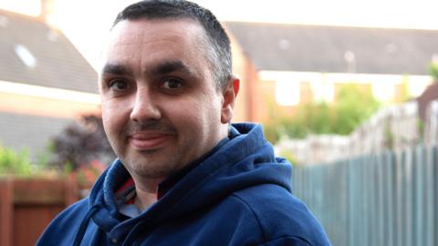 Jonathan Moran is standing in a garden which has fences around it. He is wearing a navy hooded jumper and has short black hair. There are rows of houses in the background.