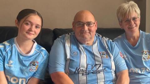 A man sits on a sofa next to a woman and a girl in a living room. All three are wearing Coventry City FC t-shirts. A dog is also sat on the floor.