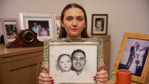 Chloe looks into the camera with a serious expression. She holds a frame which shows an old black and white picture of her as a baby with her dad. Behind her is a chest of drawers which has multiple frames displaying family pictures. 