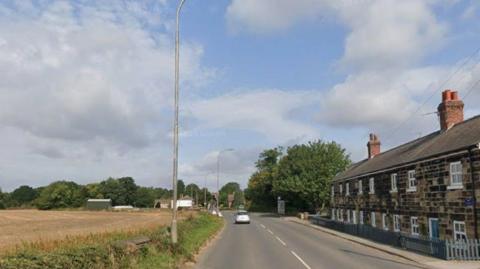 A rural road with terraced houses on one side and a farming field on the other. A single car can be seen ahead.