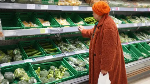 A woman, wearing a furry orange coat and orange beanie, looks at vegetables in a supermarket.