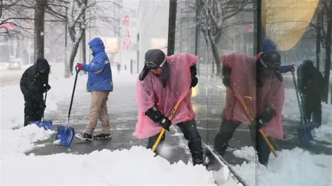 Three people shovel deep snow on a pavement while it continues to snow around them