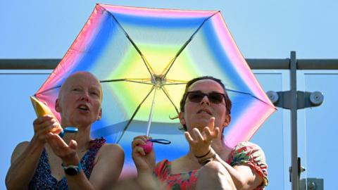 Two women sit underneath a pink, blue and yellow umbrella in the sun at Wimbledon and apply sun cream 