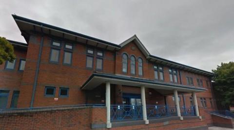 A red-brick court building which has ramp access to the left hand side and step access to the right hand side. There are blue railings, and trees either side of the building.