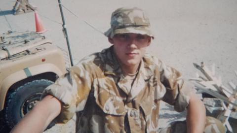 Rikki Drury pictured as a young man in combat fatigues with a camouflage hat on is standing near a four-by-four vehicle. 