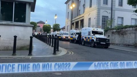 A number of police Land Rovers can be seen in a row near to Nailors Row in Londonderry. Black bollards line the other side of the road. A large pink building can be seen in the background as well.  