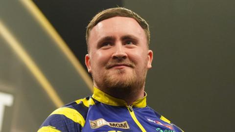 Darts player Luke Littler, in a blue and yellow top and smiling