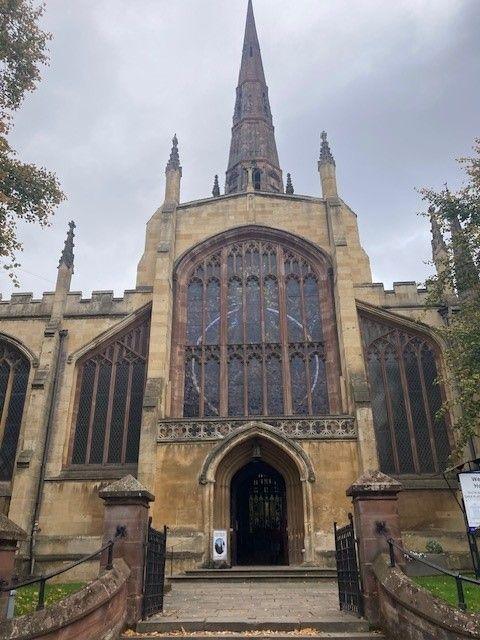 A view of a medieval sandstone church taken from the path outside. The church's entrance can be seen, large stained glass window and tall spire.