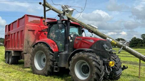 A red tractor and trailer in a farmer's field with a large electricity pole lying across the front and roof