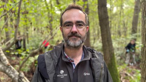 Woodland Trust site manager David Logan is pictured in the foreground wearing a black Woodland Trust fleece. Trees and people working are blurred in the background.