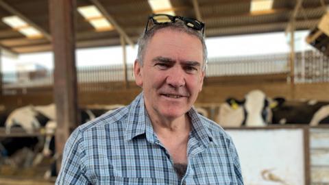 Llun o'r amaethwr a llywydd undeb NFU Cymru, Aled Jones, yn sefyll mewn cwt gwartheg gyda'r gwartherg yn pori y tu ol iddo. Mae'n gwisgo crys siec sgwarog glas ac mae ganddo sbectol haul am ei ben. Mae'r gwartheg tu ol iddo yn ddu a gwyn eu lliw. 
