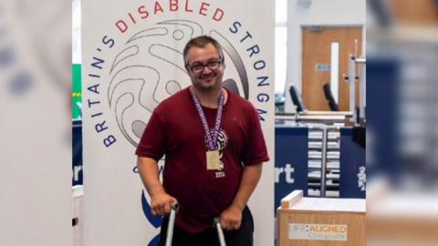 Gary Clarke wearing a burgundy t shirt and glasses with a purple lanyard. Gary is standing in front of a big sign which reads Britain's Disabled Strongman. He is standing with crutches. 