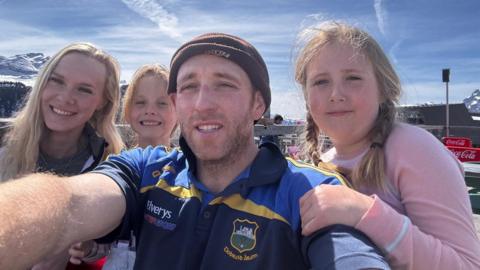 James holds out the camera for a selfie with his family of two girls and his partner Ellie. They appear to be in a ski resort as there are snowy mountain tops behind. He wears short sleeved T-shirt and beany hat.