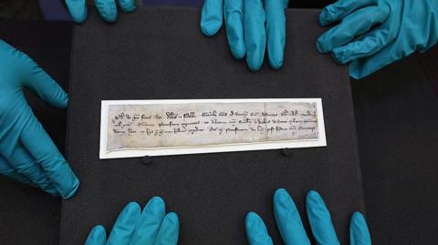 A small piece of parchment sitting on a black block surrounded by hands with wearing blue rubber gloves. 