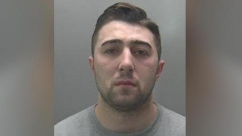 Custody image of Jordan Neale. He has short cropped dark hair and dark stubble, and is wearing a grey jumper.