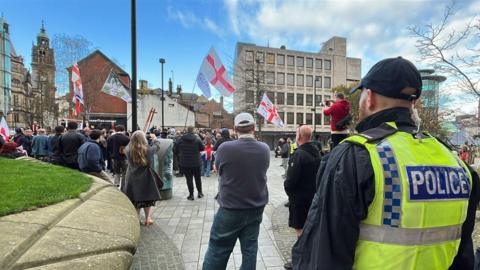 A public gathering in an urban square with several people standing and facing a group holding flags featuring the red cross of St George. Two police officers in high-visibility jackets are in the foreground, observing the scene.