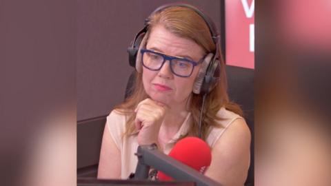 A woman with shoulder-length light ginger hair sits in front of a microphone and looks at someone off-screen