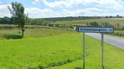 A grassy verge on a countryside road. There is a sign that is pointing left that says 'Kirkwhelpington 1/4'. It is a bright summers day, with blue skies and some white clouds. There are also some wildflowers, and lots of green trees.