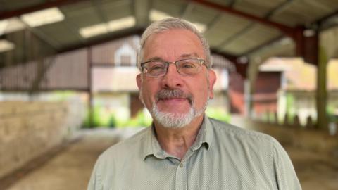 Guy Hall who has grey hair, beard and glasses standing in an old barn in Stanmer Park which he hopes his charity can use as a workshop and museum