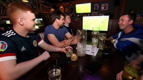 Four Scotland fans sitting in a pub watching their team play. Two of the men are wearing Scotland football shirts , another has the Saltire flag wrapped around him.