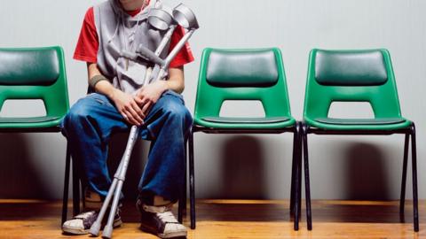 A patient in a t-shirt and jeans sits holding a pair of crutches and waits in a waiting room