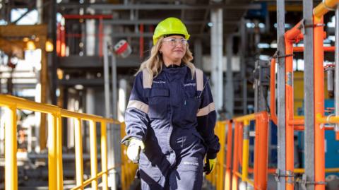 A woman in working gear and a high vis helmet is walking across a platform at a building site. 