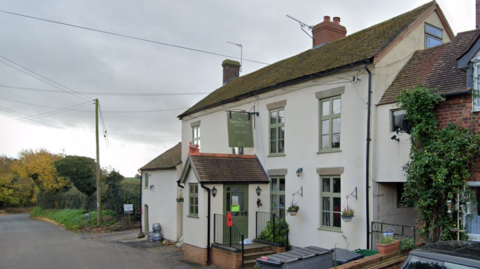 A white pub building with green moss on the roof. It has green window panes and a green sign and door. There is a small enclosed porch and a poppy on a metal rail outside