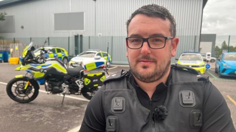 Roads Policing Inspector Matt Boiles from Avon and Somerset Police stands in a car park with police cars and motorbike behind him.