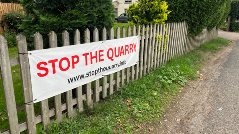A white banner with the words Stop The Quarry written on it in red. It is attached to a wooden fence. 