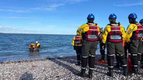 Coastguard recruits wearing dry suits to the right watching three people in the water. They are stood at Brixham Beach.