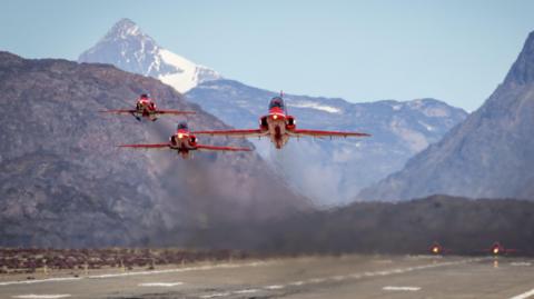 Three Red Arrows aircraft seen taking off towards the camera. They are just above the runway with partially snow-covered mountains in the background.