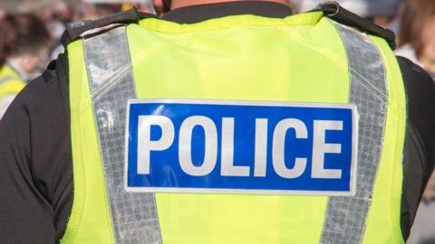 Close-up of police officer's back. He is wearing a hi-vis jacket with the word Police in white letters on blue.