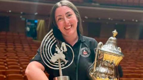A woman called Stephanie Binns holds two silver trophies in a concert hall she won with The Flowers Band. She is smiling. She is wearing a black polo shirt with her band's logo on it. Binns has long brown straight hair.