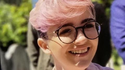 A woman with short pink hair wearing black glasses and smiling.