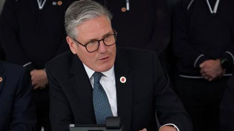 Sir Keir Starmer smiles while speaking on the phone to President Trump from a car factory in the West Midlands.  He is wearing a dark colour suit with a blue patterned tie and has a poppy lapel pin to signify the 80th anniversary of VE Day. 