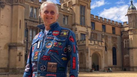 A man with grey hair is stood outside Knebworth house wearing a blue denim jacket covered in band badges