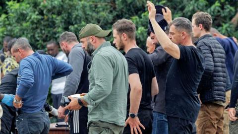 Romanian mercenaries - not in uniform - pictured side on in the rain being checked by Rwandan security guards at the border, one of the men has his hands up - Wednesday 29 January 2025