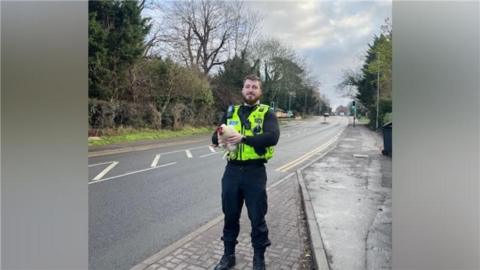 A police officer stands to the side of an empty road holding a white chicken. 