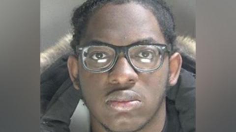 A police custody image of Sakeen Gordon, wearing black-rimmed glasses. He is looking straight at the camera. He is wearing a black coat with a fur trim. He is not smiling. 