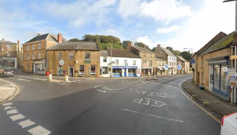 A road with market square in Crewkerne. It has a mini-roundabout and shops in the background.