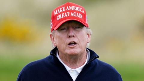 Donald Trump looking slightly off camera. He is only visible from the shoulders up. He has a red cap on his head with the words: 'Make America Great Again' in white writing on the front. He is wearing a white polo shirt under a dark blue zip-up jumper.