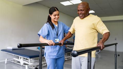 A female physiotherapist with long black hair wearing blue medical scrubs helps a man in a yellow shirt who is holding on to parallel bars 