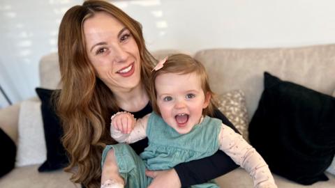 A young woman with long auburn wavy hair smiles into the camera with a baby girl sitting on her lap. The child is also smiling at the camera.