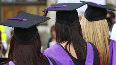 The backs of three girls in black and purple graduation caps and gowns. Two have long dark hair the other has long blonde hair.