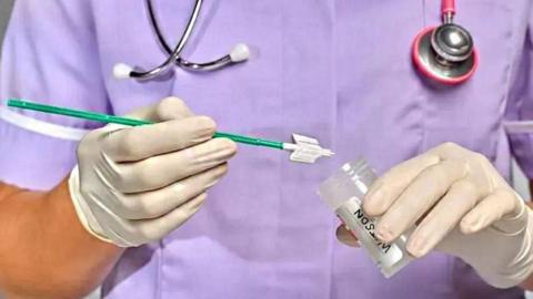 a close up of a nurse wearing a lilac uniform and surgical gloves preparing a smear test for cervical screening