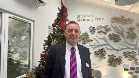 George Lillie wearing a suit jacket with a pink and grey striped tie. There is a Christmas tree behind him to the left and wall décor on the right.