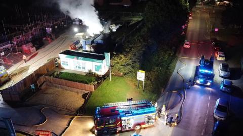 An aerial view shows fire engines at the scene which is lit up in the dark of the early morning. Smoke is billowing from the top of the container